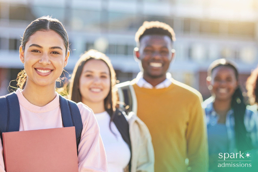 Diverse group of smiling college students standing outdoors on campus, with one student holding a folder in the foreground.