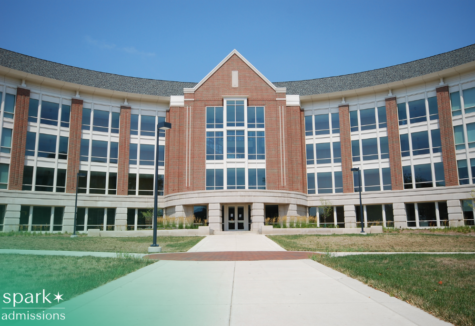 Large brick academic building with a curved facade and tall glass windows, viewed from a walkway leading to the main entrance on a clear day.