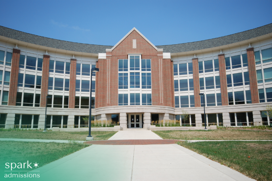 Large brick academic building with a curved facade and tall glass windows, viewed from a walkway leading to the main entrance on a clear day.