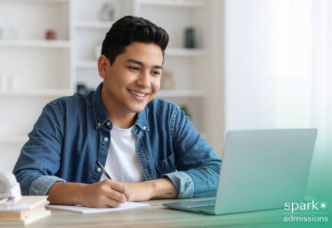 High-achieving student with a learning difference smiles while taking notes and working on a laptop at a desk in a bright home study space.
