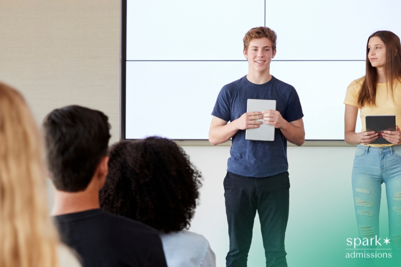 Two students stand at the front of a classroom giving a presentation, holding tablets and speaking to seated classmates.