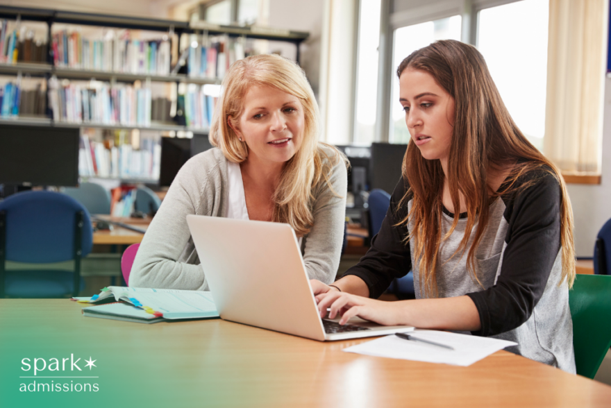 Tutor or teacher helps a student with laptop work at a library table, reviewing notes and assignments during an academic support session.