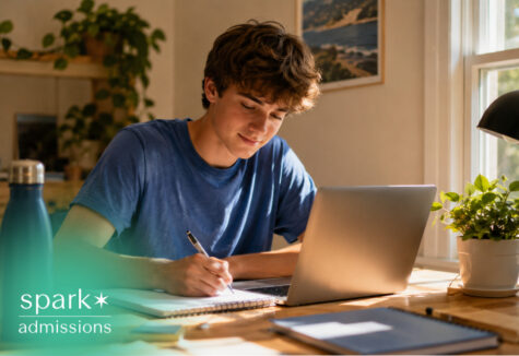 Student writing notes beside a laptop in a sunny home study space